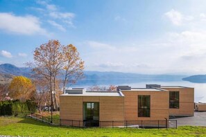 A modern building with a flat roof, large windows, and a view of a lake and mountains in the background. - WE Hotel Toya - Dusit Collection (Toyako)