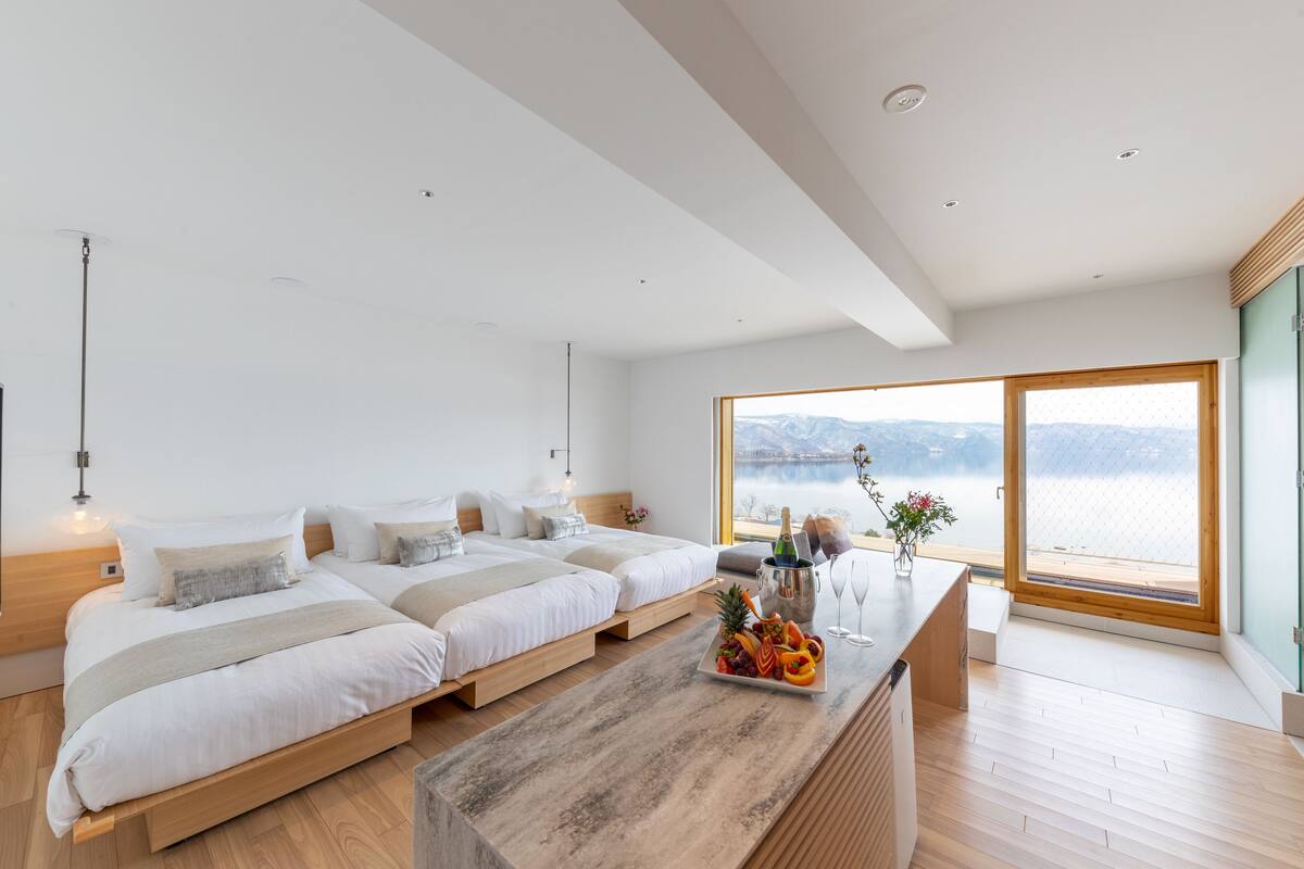 Ett modernt hotellrum med ett stort fönster som erbjuder utsikt över en sjö och berg. Det finns två sängar med träramar och vita sängkläder. Ett träbord med frukt och en flaska champagne står i mitten av rummet.