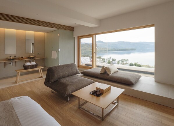 A modern room with a sofa, a small table, a bench, and a large window offering a view of a lake and mountains. - WE Hotel Toya - Dusit Collection (Toyako)