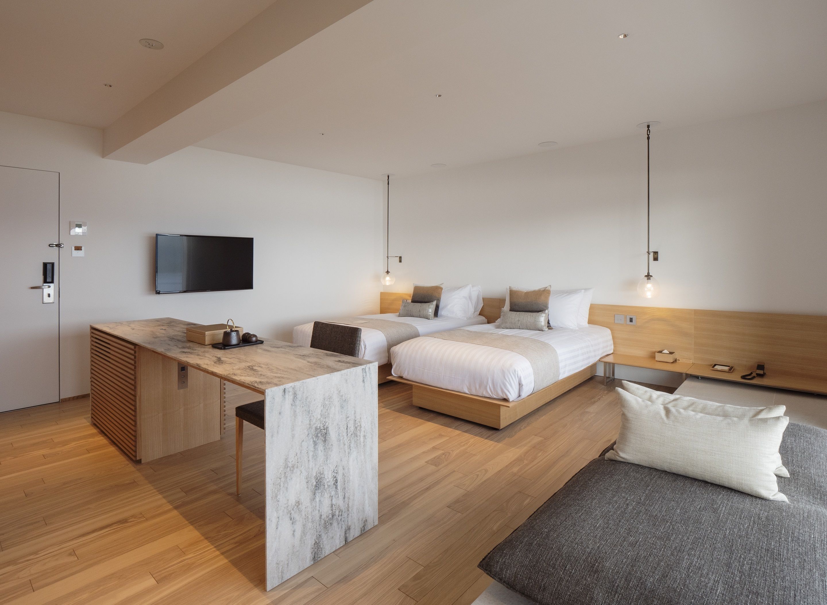 A modern hotel room with a wooden floor, a desk with a marble top, two beds with white bedding, a grey sofa, and a flat-screen TV mounted on the wall.