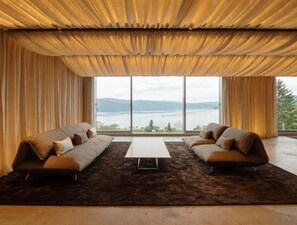 A modern living room with a large window, two sofas, a coffee table, and a view of a lake and mountains. - WE Hotel Toya - Dusit Collection (Toyako)