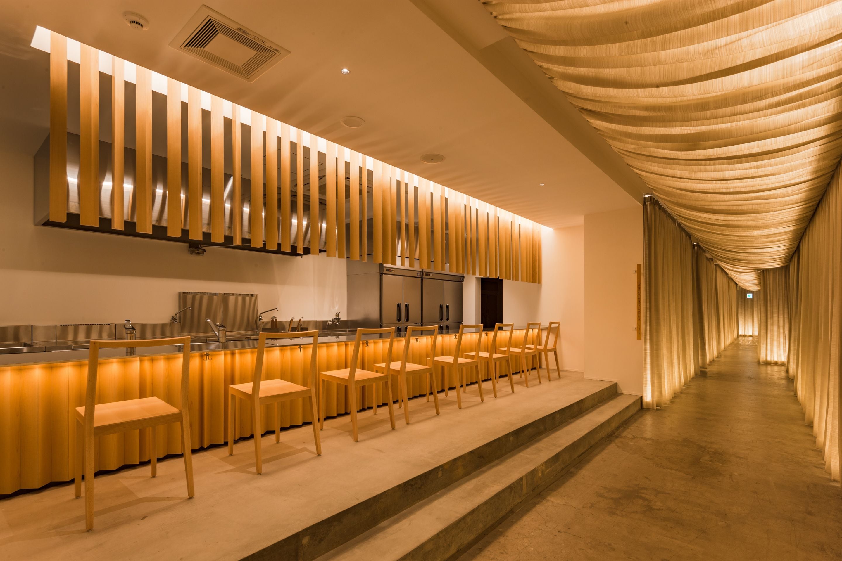 A modern kitchen with a long counter, wooden chairs, and a unique ceiling design.