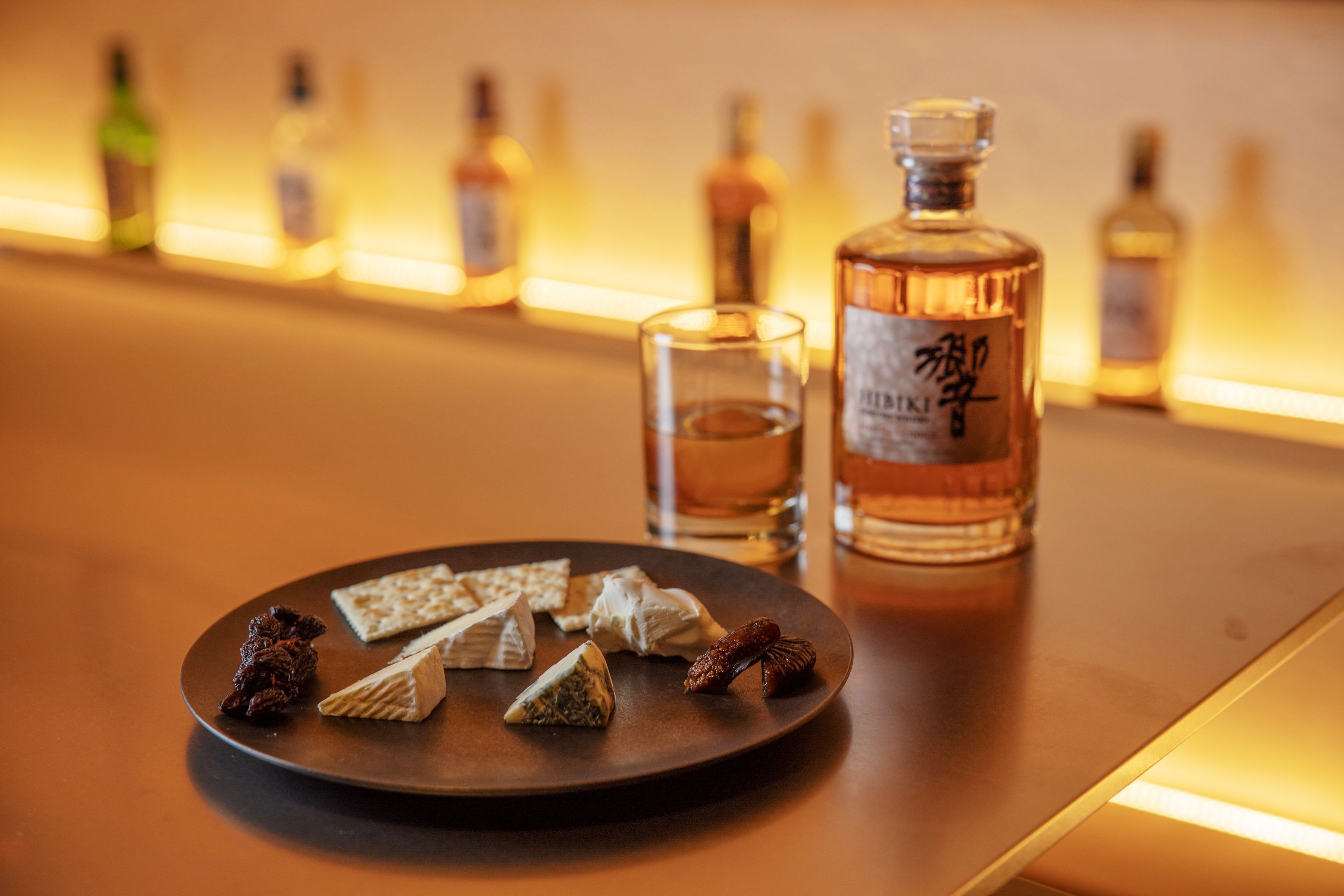 A bar setting with a bottle of whiskey, glasses, and assorted snacks on a plate.