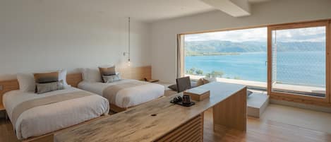 A modern bedroom with two beds, a wooden desk, and a large window offering a view of a lake and mountains.