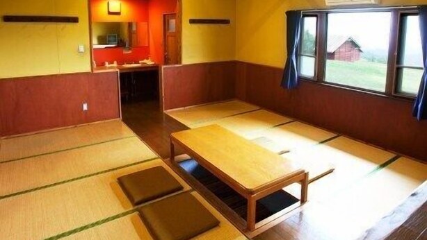 A tatami mat room with a wooden table, a window with curtains, and a small kitchen area. - Kujukogen Cottage (Taketa)