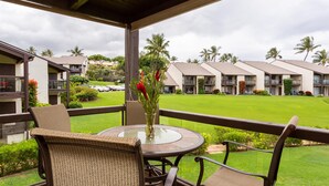 A covered outdoor seating area with wicker chairs and a glass table, overlooking a well-maintained lawn and a row of hotel buildings.