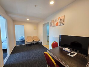 A hotel room with a bed, two chairs, a TV, and a desk with a remote control. - Empire Apartments (Auckland)