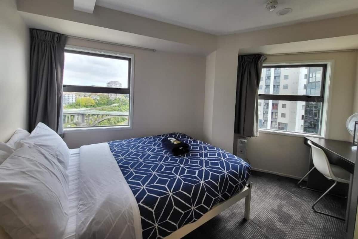 A hotel room with a bed, a desk, and a view of buildings through the window.