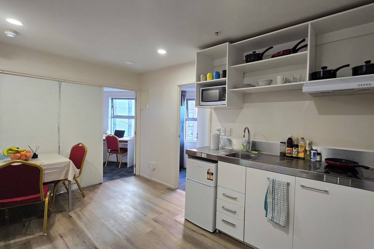 A compact kitchen with white cabinets, a microwave, and a sink. There's a dining area with a table and chairs, and a view into a room with a desk and chair.