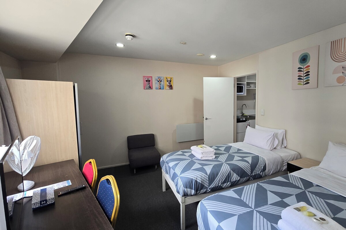 Habitación de hotel con dos camas, un escritorio con silla, armario y kitchenette.