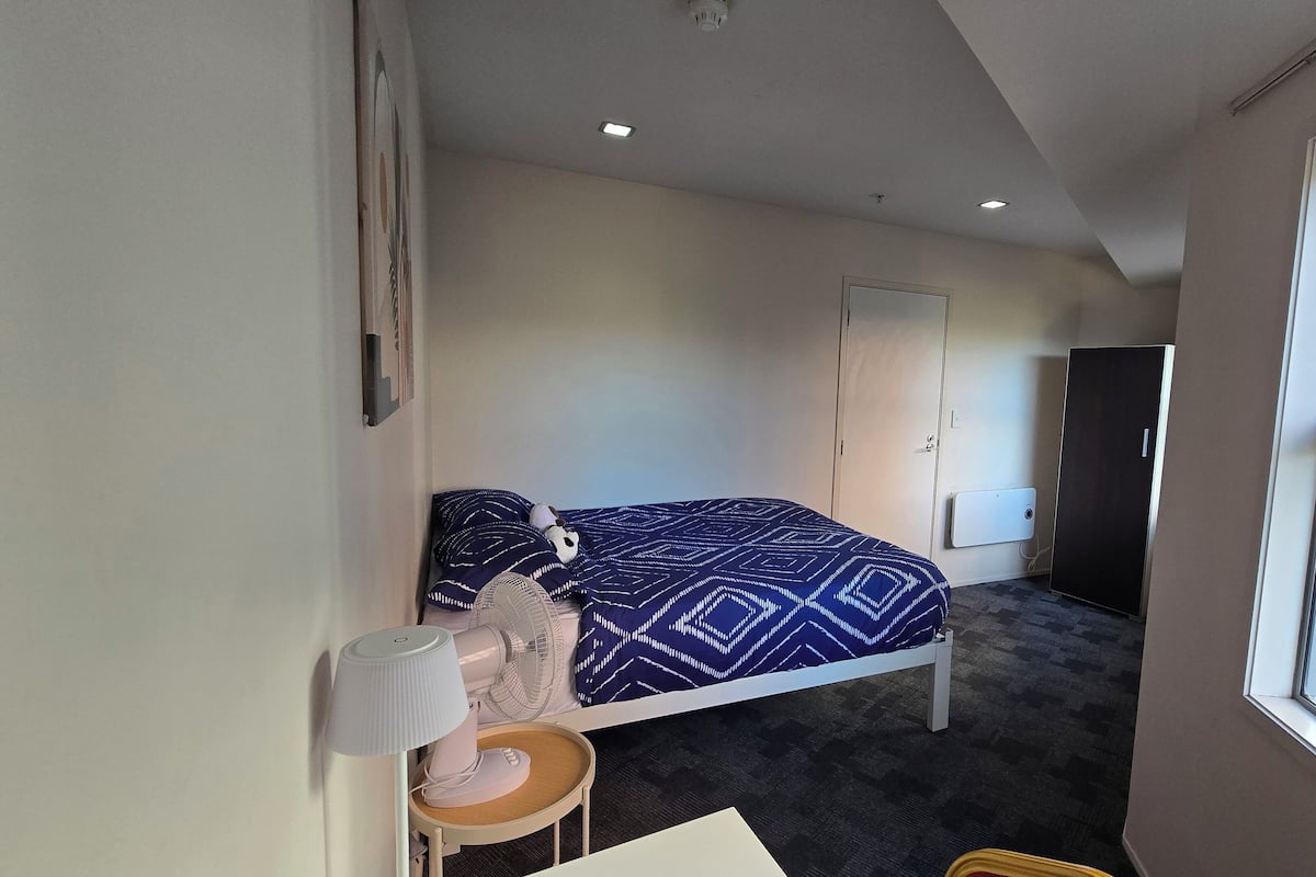 A bedroom with a bed, a bedside table with a lamp, a small stool, a wardrobe, and a window with a view of the street.