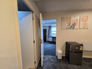 A hotel room with a door, a refrigerator, a trash bin, and a desk with a chair. - Empire Apartments (Auckland)