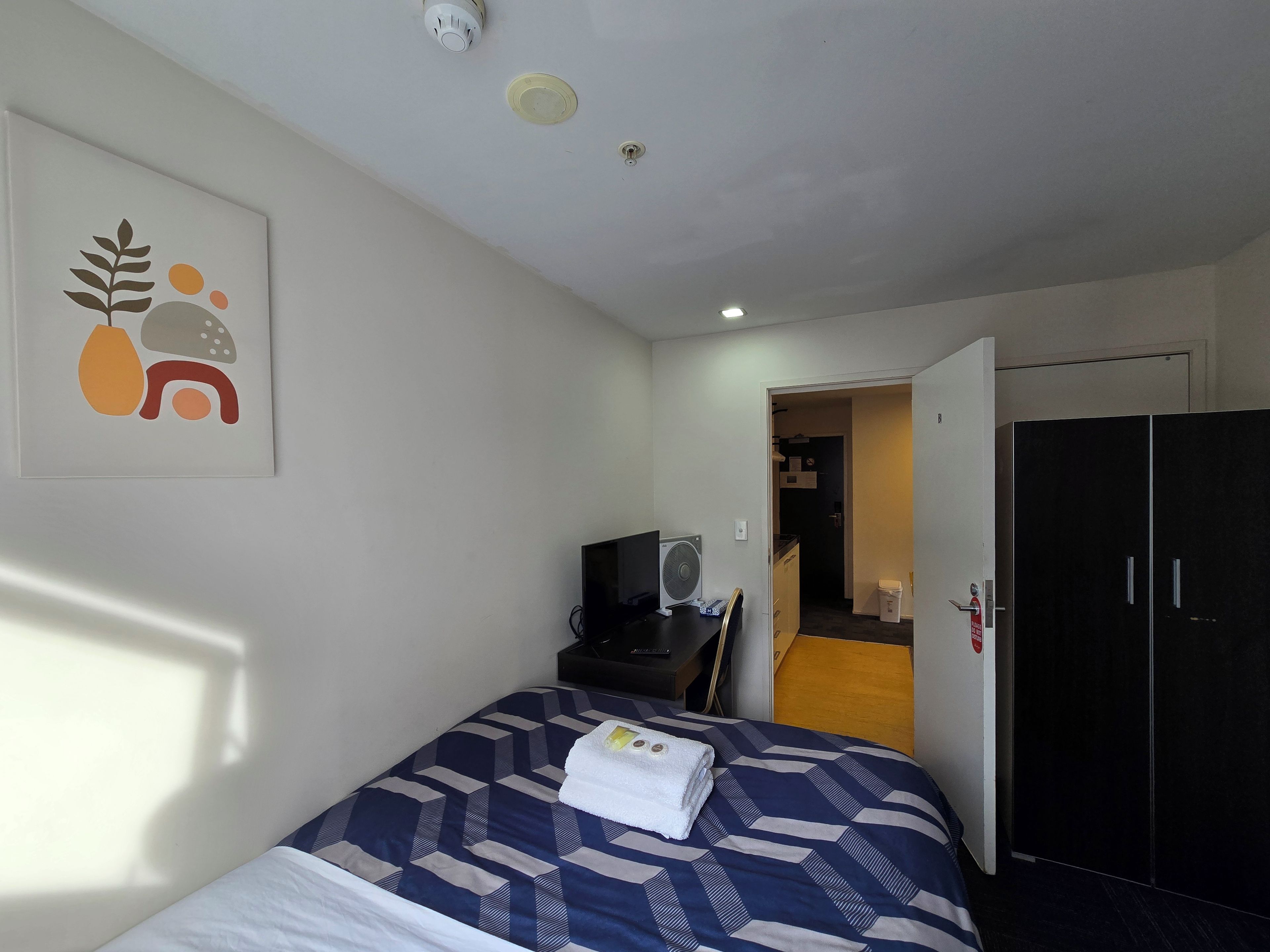 Ein Hotelzimmer mit einem Bett, einem Schreibtisch mit Computer, einem Ventilator und einer Tür zu einem Flur.