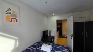 Ein Hotelzimmer mit einem Bett, einem Schreibtisch mit Computer, einem Ventilator und einer Tür zu einem Flur.