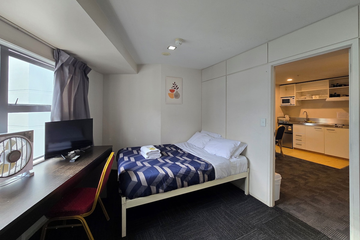 Una habitación de hotel con una cama, un escritorio con una silla, un televisor, un ventilador y una puerta abierta que da a una kitchenette.