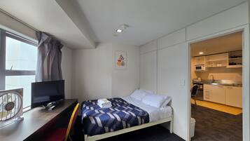 A hotel room with a bed, a desk with a chair, a TV, a fan, and an open door to a kitchenette.