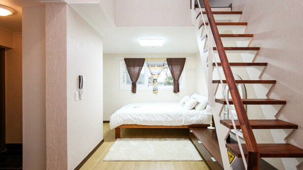 A bedroom with a wooden bed, a white rug, a staircase, and a window with curtains. - Beanfield House (Seogwipo)