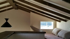 A loft-style bedroom with exposed wooden beams, a slanted ceiling, a window with a view, a bed with checkered bedding, and a black pendant light. - Beanfield House (Seogwipo)