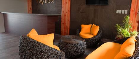 A modern lounge area with wicker furniture and orange cushions, a wooden reception desk, and a wall-mounted TV.