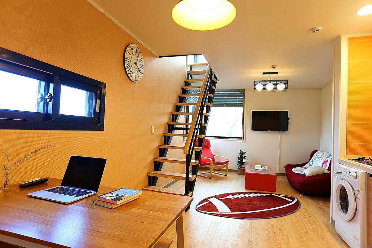 A modern living room with a wooden staircase, a red couch, a washing machine, and a desk with a laptop and books.