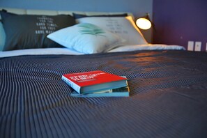 Una cama bien hecha con un libro rojo encima, una almohada con un diseño verde y una lámpara fijada a la pared.