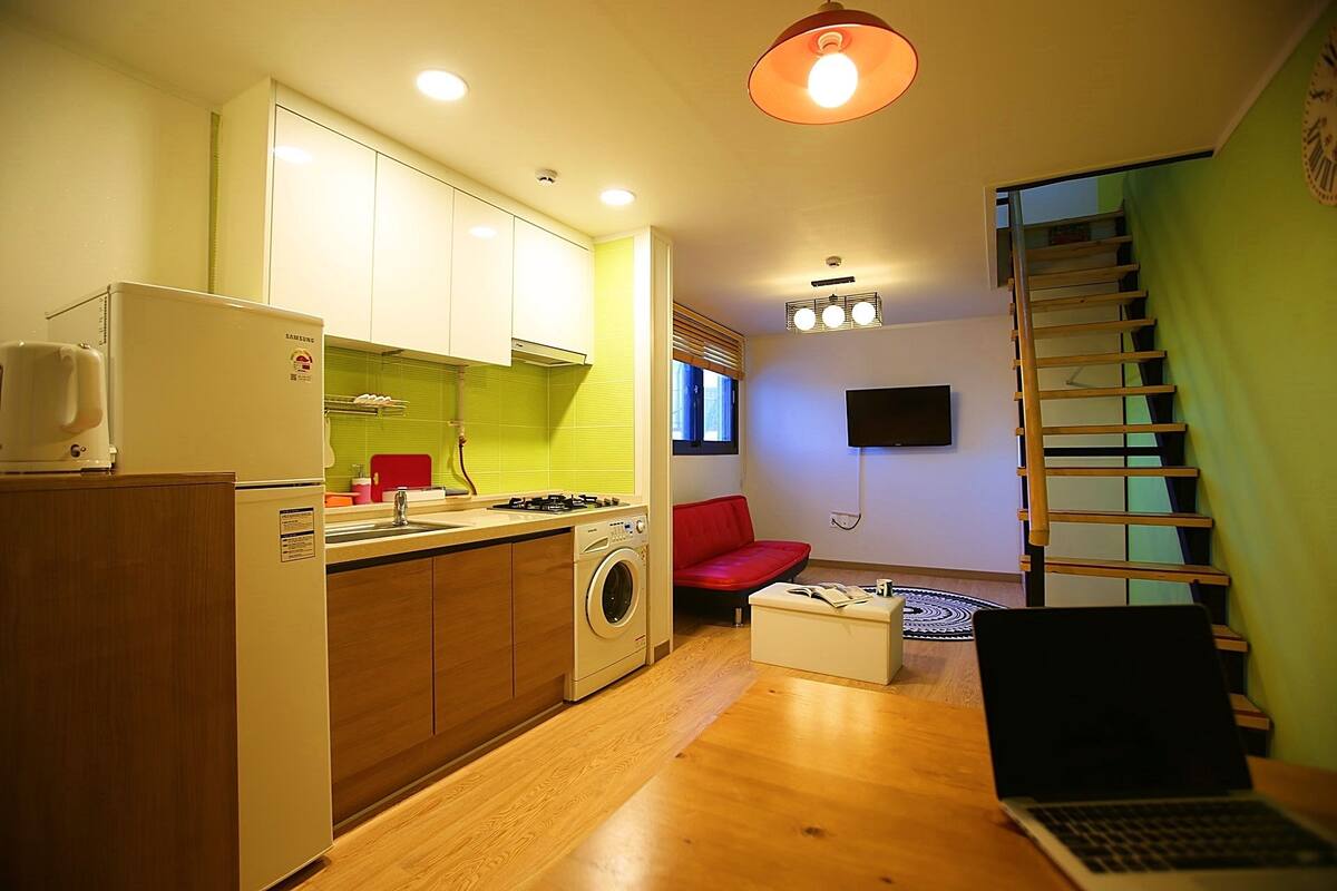 A compact kitchen with a washing machine, a red sofa, a laptop, and a TV.