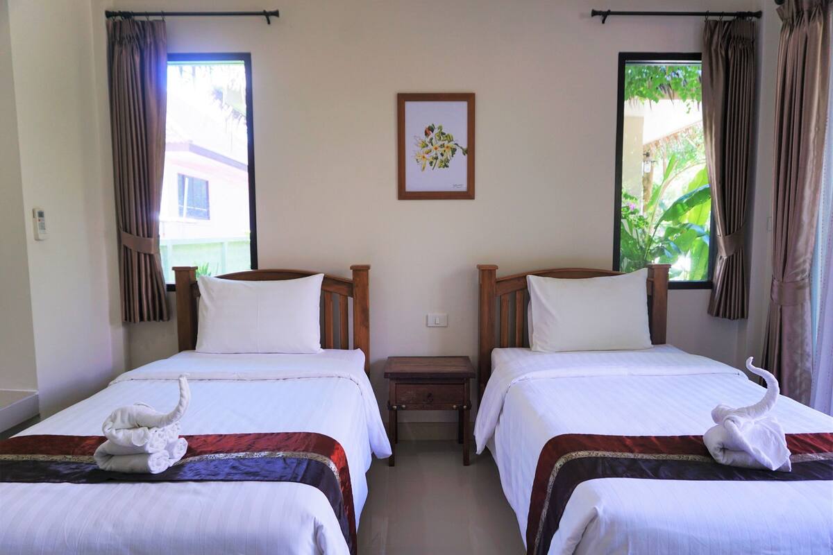 A hotel room with two single beds, each with a wooden headboard, white bedding, and a swan-shaped towel on the bed. There is a small bedside table between the beds, and a framed picture on the wall above the beds.