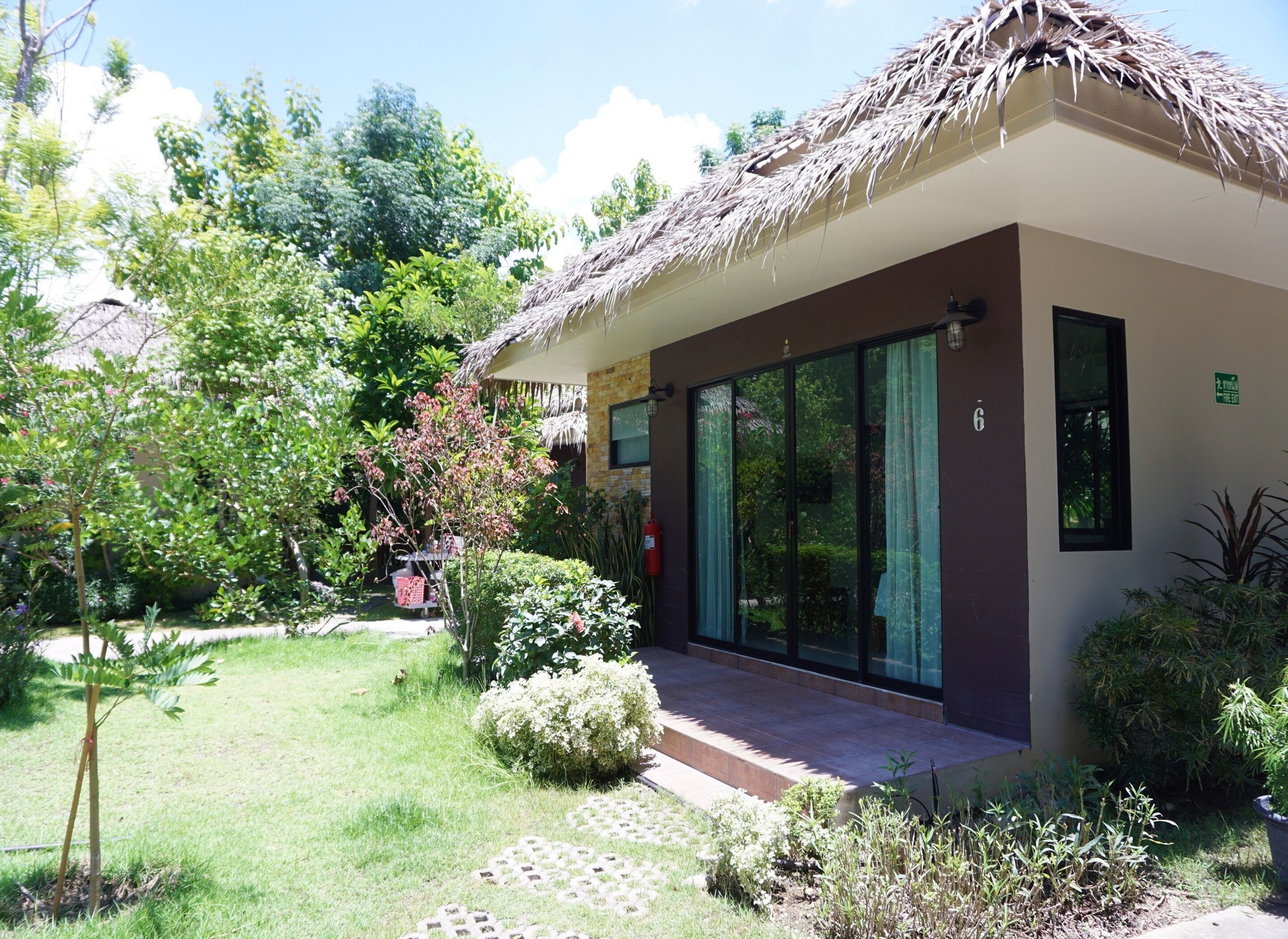 A modern building with a thatched roof, large glass doors, and a well-maintained garden with various plants and a small tree.