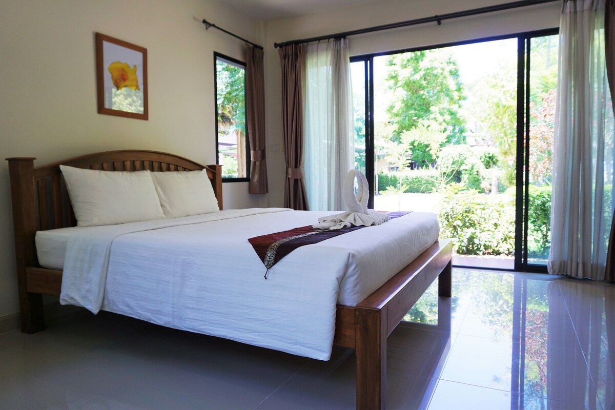 A neatly made bed with white linens and a decorative pillow, a wooden headboard, and a view of greenery through large windows.