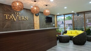 A reception area with a wooden reception desk, a TV mounted on the wall, and a seating area with a yellow sofa and round black chairs. - The Tavern Villas at  Orchid Gardens (Bacolor)
