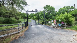 A gated entrance with a metal gate, a wooden fence, and a road leading into a green area with trees.