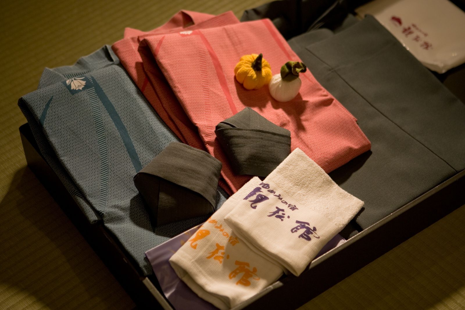 Prendas de ropa ordenadamente dobladas, incluyendo una camisa gris estampada, una camisa roja y una camisa marrón, junto con una toalla blanca con escritura japonesa.