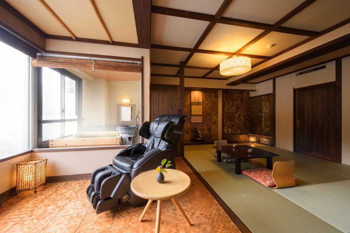 Een kamer met tatami vloerbedekking, een massagestoel, een klein rond tafeltje met een vaas en een traditionele Japanse tafel met kussens.