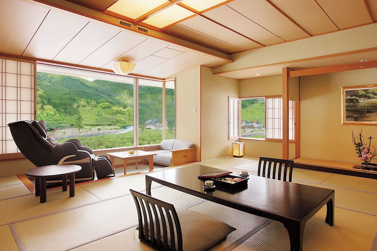 Een traditionele Japanse kamer met tatami vloerbedekking, een lage tafel, stoelen, een bank en grote ramen met uitzicht op een bebost gebied.