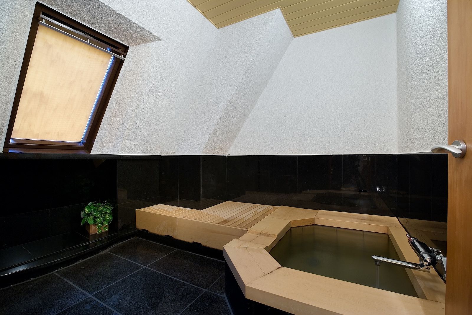 A modern bathroom with a freestanding bathtub, black tiled walls, and a skylight.