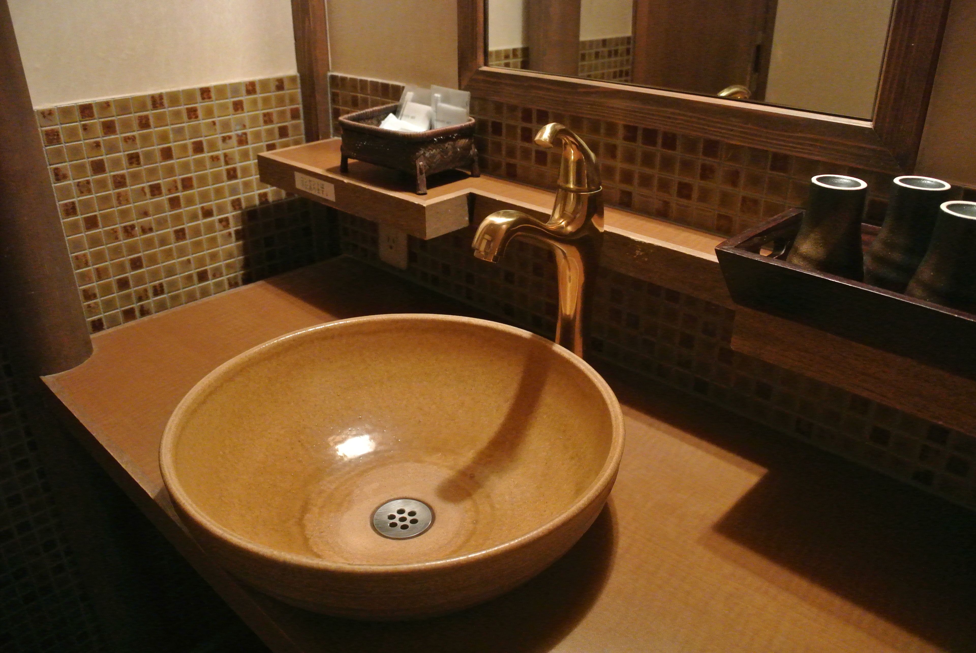 A bathroom with a round ceramic sink, a gold faucet, a small shelf with a tissue box, and a mirror above the sink.
