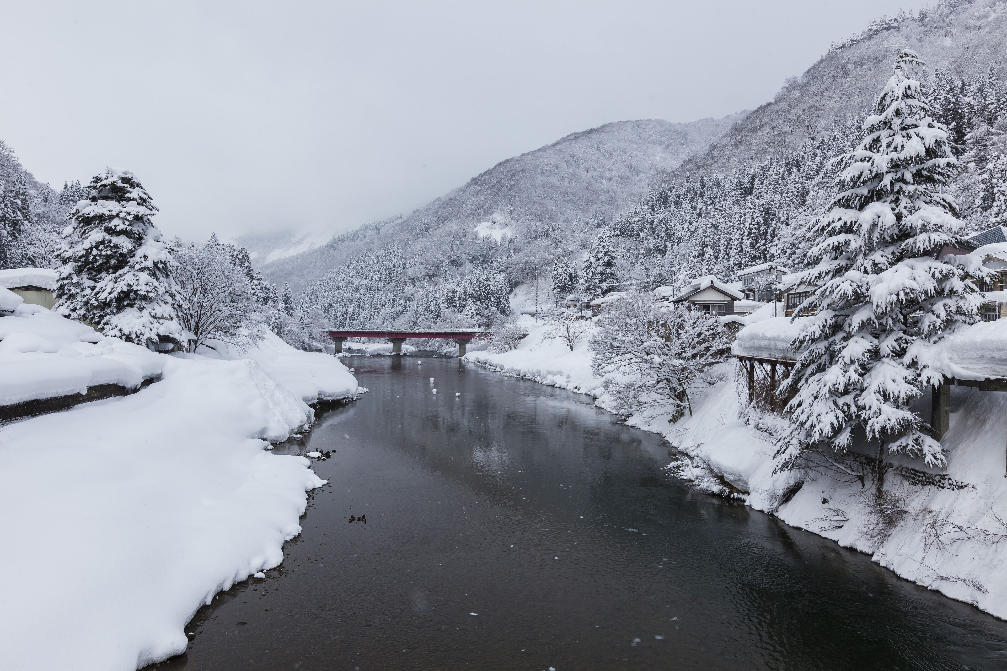 A snow-covered river with a bridge, surrounded by snow-laden trees and mountains.