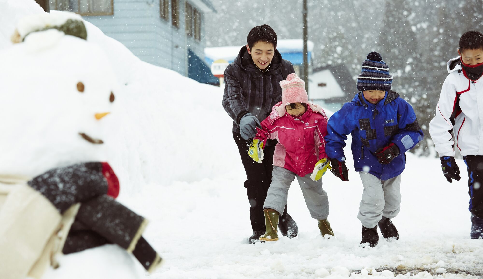 Un muñeco de nieve con nariz de zanahoria y ojos de carbón, una persona con una chaqueta negra y dos niños vestidos para el invierno.
