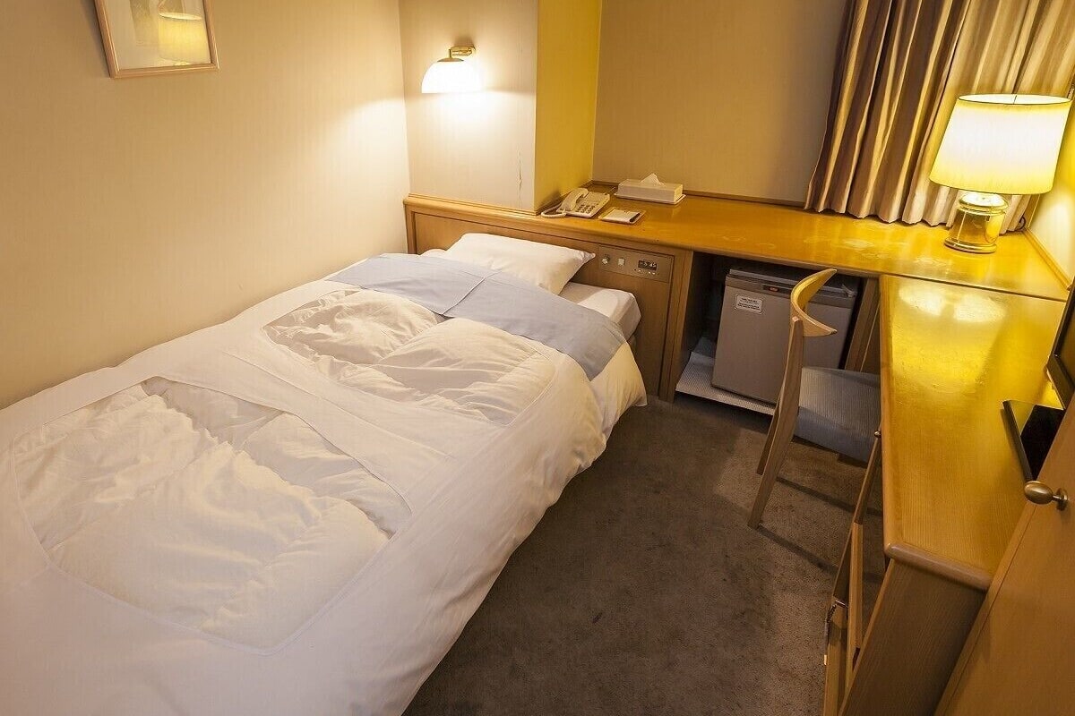 Een enkel bed met witte lakens, een nachtkastje met lamp, een telefoon en een klein bureau met een stoel.