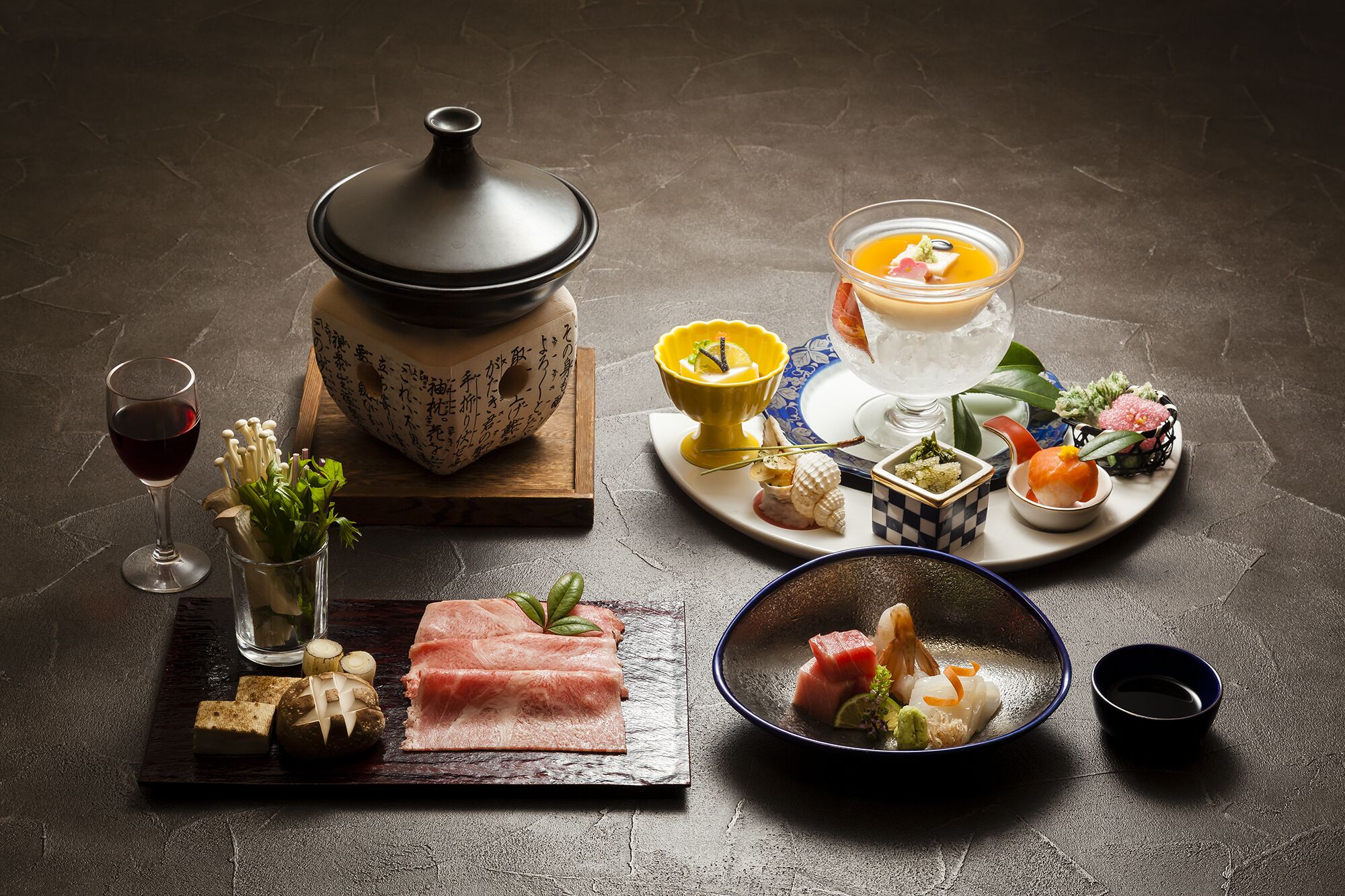 A variety of Japanese dishes including sashimi, sushi, and a hot pot, arranged on a dark textured table.