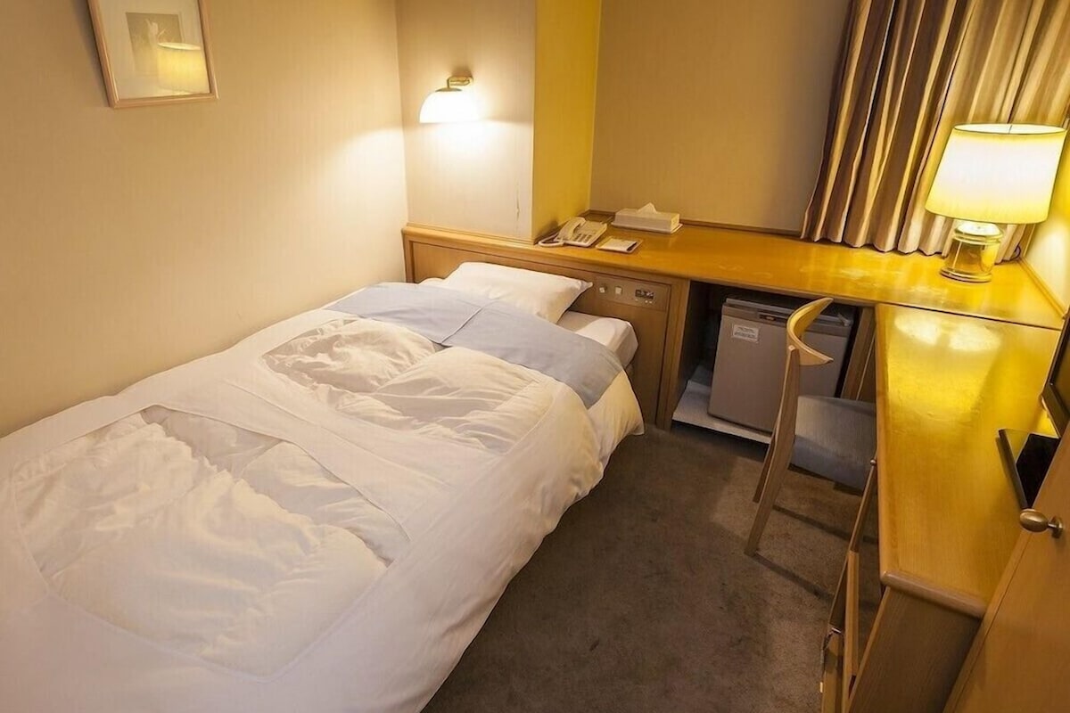 A hotel room with a bed, bedside table, lamp, and a small desk with a telephone and papers.