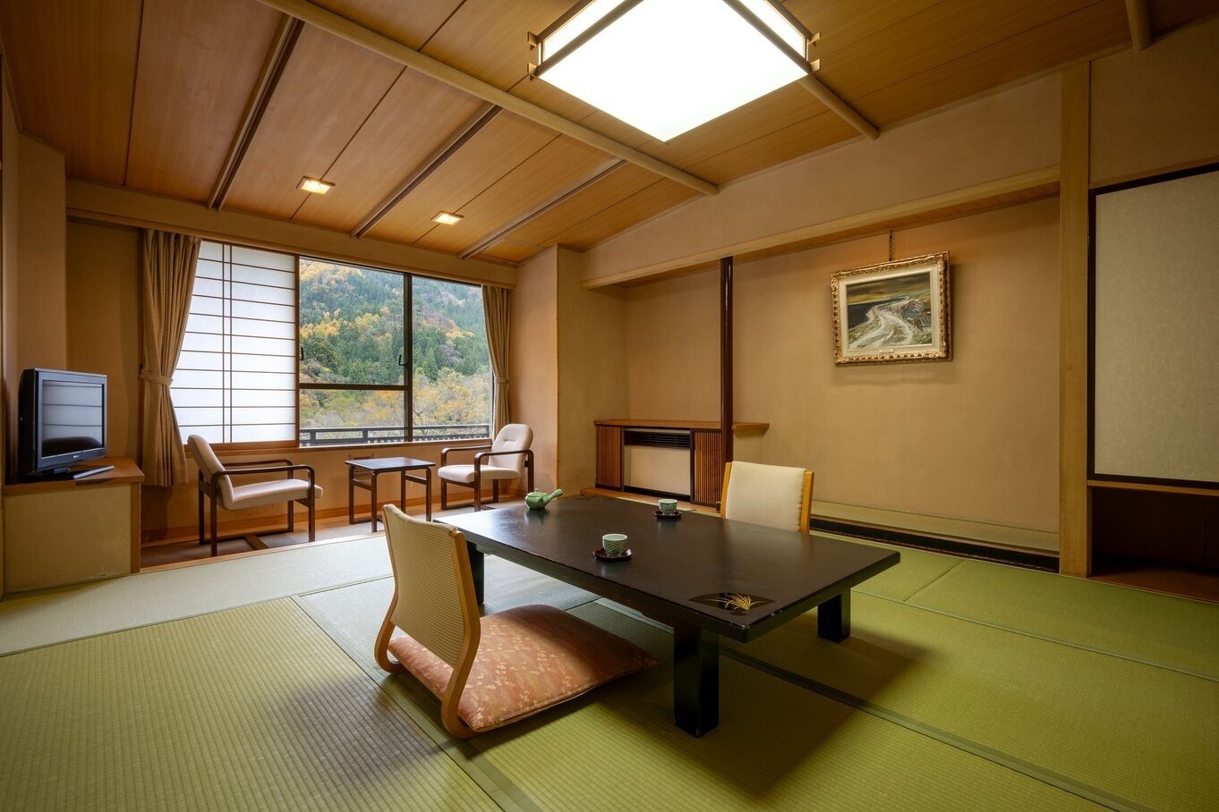Een traditionele Japanse kamer met tatami vloerbedekking, een lage tafel, stoelen, een televisie en een schilderij aan de muur.
