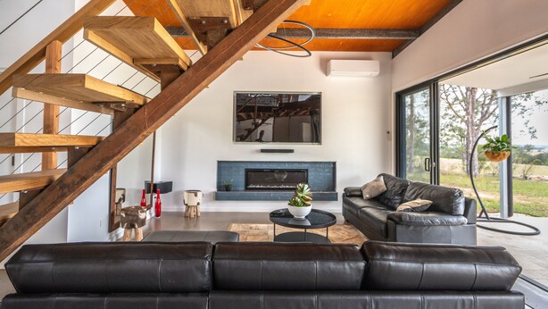 Un salon moderne avec un canapé en cuir noir, un escalier en bois, une cheminée et de grandes fenêtres.