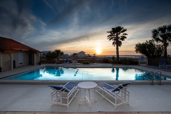 A swimming pool with lounge chairs and a table, surrounded by palm trees and a sunset view.