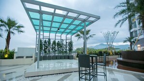 Un área de estar al aire libre con techo de cristal, rodeada de palmeras y montañas al fondo.