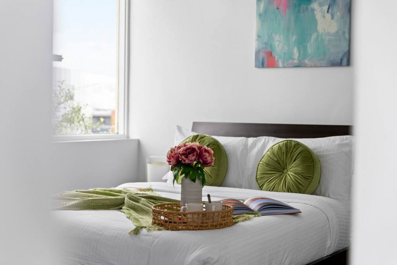 A neatly made bed with a green throw blanket, a vase with flowers, and an open book on the bedside table.