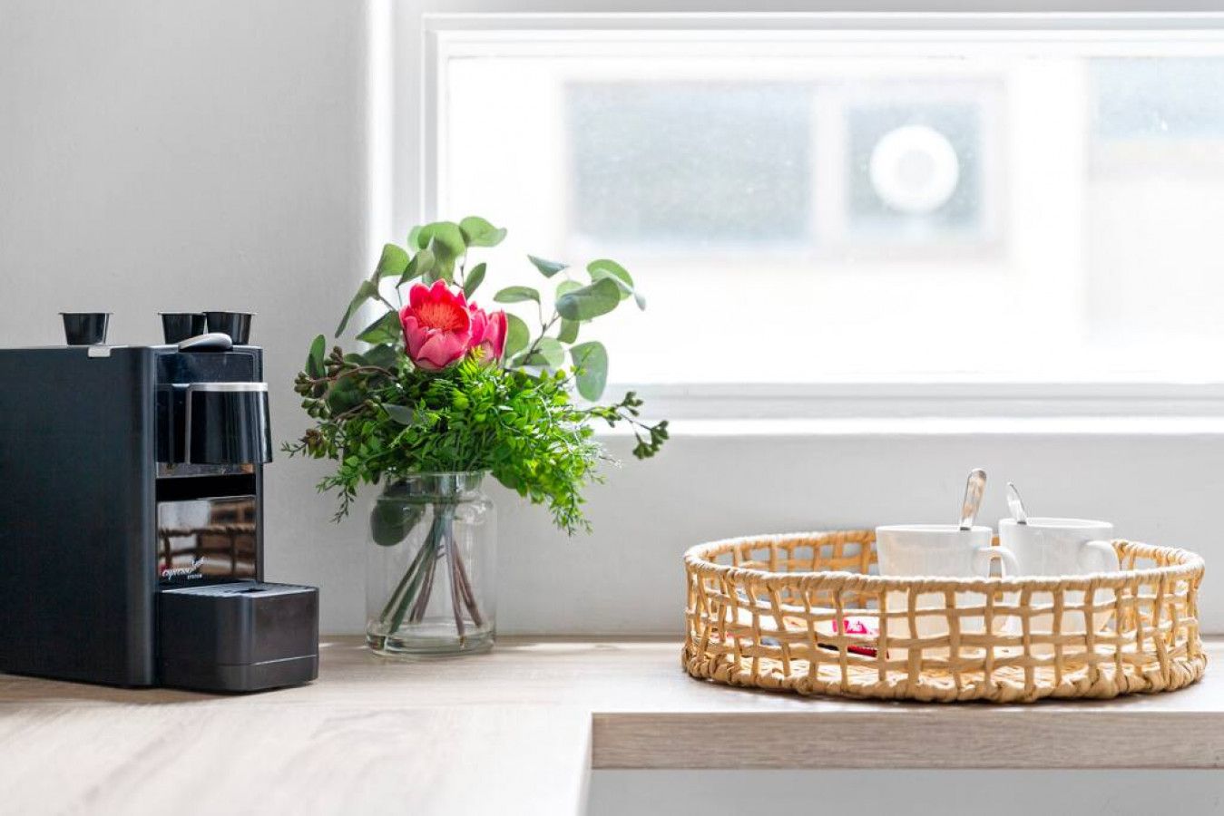A coffee machine, a vase with a flower, and a basket with cups on a wooden surface.
