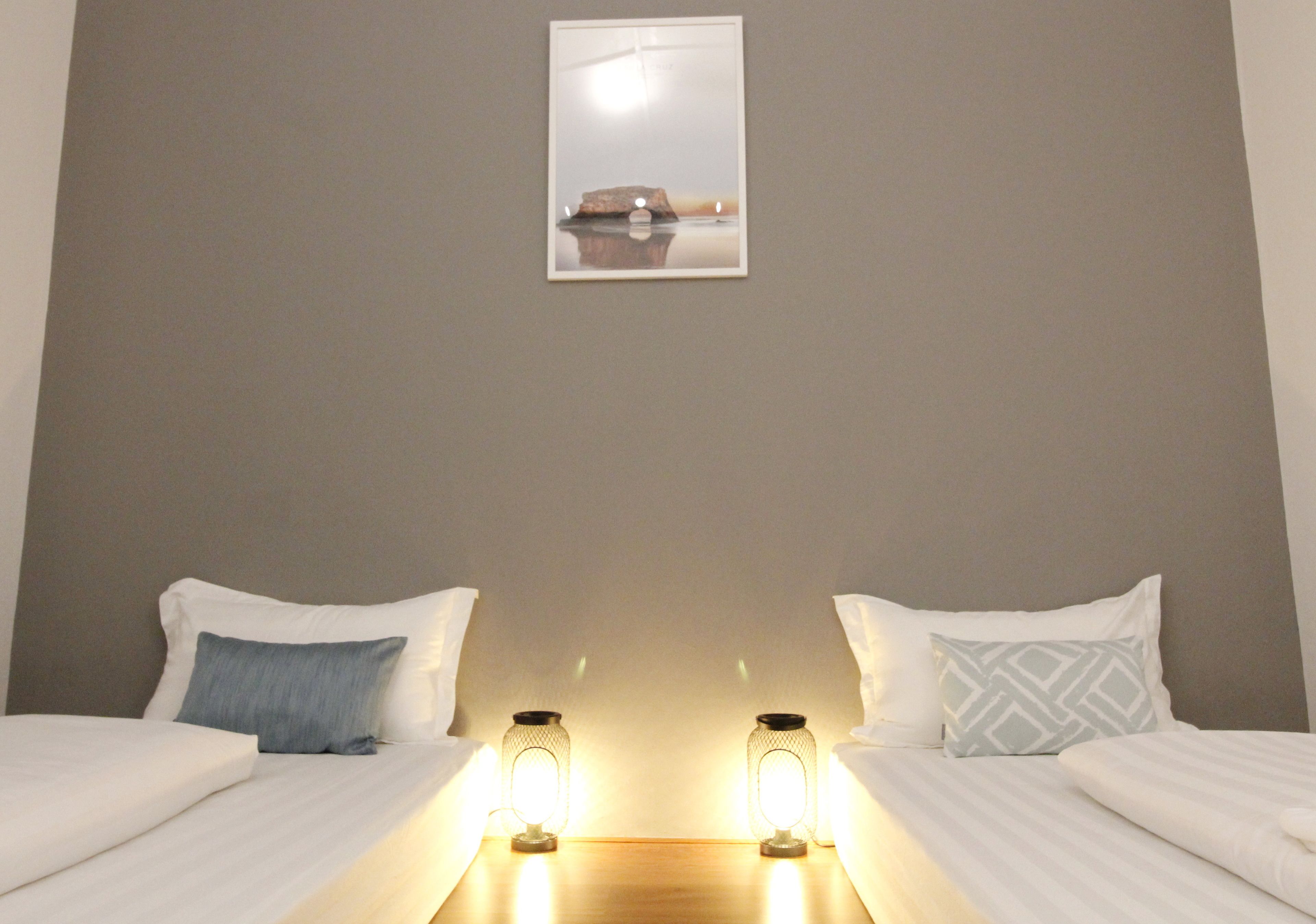 A hotel room with two beds, each with white bedding and a single pillow. There are two wall-mounted lamps on the nightstands.