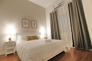 Una cama bien hecha con sábanas blancas y dos almohadas, una mesita de noche con lámpara y un aire acondicionado de pared.