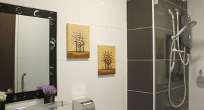 Un baño con un espejo grande, dos cuadros colgados en la pared, una ducha y un dispensador de papel higiénico.